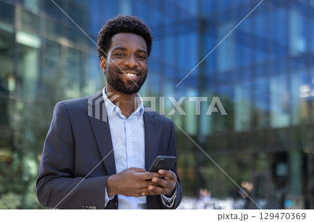 Confident businessman standing outdoors in front of a modern glass building using a smartphone. Dressed in formal wear, he exudes professionalism while engaging with his mobile device. 129470369