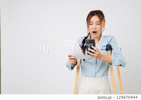 Injured woman with crutches holds mobile phone and bill paper, confused by calculator numbers. Crutches help her stand as she struggles with payment problems. isolated studio white background Injured woman with crutches holds mobile phone and bill paper, confused by calculator numbers. Crutches help her stand as she struggles with payment problems. isolated studio white background 129470444