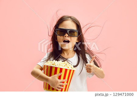 Portrait of Cute Cheerful Child Girl in 3D Glasses, holding popcorn bucket, Showing ThumbUp, Expressing Surprised, Wearing in Classical White T Shirt Standing on Pink Isolated Background. 129471000