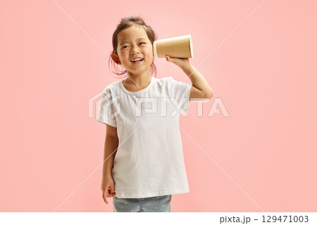 Cheerful cute Asian child girl spying using paper cup for listening, trying overheal someone, holds hand with cup near ear to enhance the sound, wearing in neutral clean white t shirt on pink isolated Cheerful cute Asian child girl spying using paper cup for listening, trying overheal someone, holds hand with cup near ear to enhance the sound, wearing in neutral clean white t shirt on pink isolated 129471003