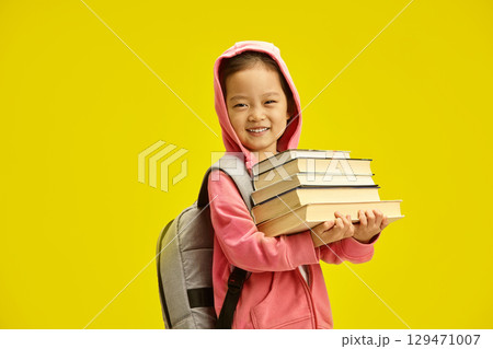 Active excellent little schoolgirl holding books going to school wears in hoodie and backpack on shoulders, isolated on yellow background. Go to school Active excellent little schoolgirl holding books going to school wears in hoodie and backpack on shoulders, isolated on yellow background. Go to school 129471007