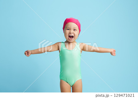 Little female child swimmer cheerfully spreads her arms to the sides, dressed in a turquoise swimsuit and a pink cap standing on a blue isolated Little female child swimmer cheerfully spreads her arms to the sides, dressed in a turquoise swimsuit and a pink cap standing on a blue isolated 129471021