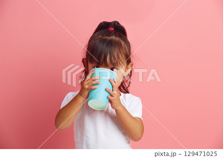 Headshot portrait of three years old Asian ethnicity little girl drinks water or juice from disposable blue paper cup standing on a pink isolated background. Headshot portrait of three years old Asian ethnicity little girl drinks water or juice from disposable blue paper cup standing on a pink isolated background. 129471025
