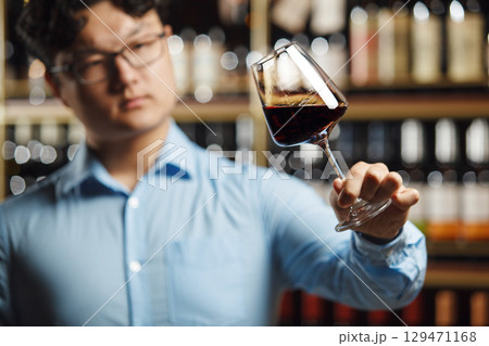 Asian Sommelier Carefully Evaluating Red Wine In Well-Stocked Cellar 129471168