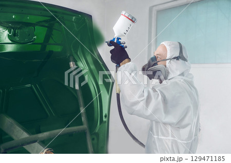 Worker in protective mask and jumpsuit overall spraying base coat of paint on car body elements in painted chamber using professional airbrush. 129471185