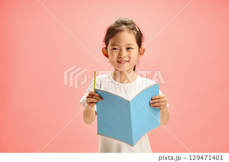 Happy and Pretty Preschool-age Korean Girl with an open Notebook with Cover Facing out and Holding Pen, Smiling Sweetly Standing on a Pink Isolated Background. Concept of Remote Learning. Happy and Pretty Preschool-age Korean Girl with an open Notebook with Cover Facing out and Holding Pen, Smiling Sweetly Standing on a Pink Isolated Background. Concept of Remote Learning. 129471401