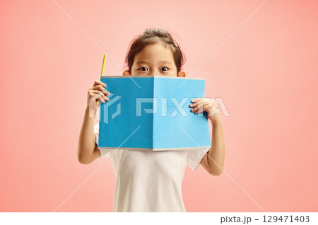 Curious Elementary Age Child Girl with Big Eyes Looks Out from Behind an open Blue Notebook with Cover on Outside, Expresses Surprised and Interest in Knowledge Standing Over Pink Isolated Background Curious Elementary Age Child Girl with Big Eyes Looks Out from Behind an open Blue Notebook with Cover on Outside, Expresses Surprised and Interest in Knowledge Standing Over Pink Isolated Background 129471403