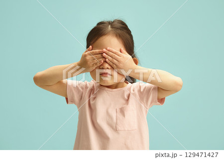 Kid girl with serious face covering her eyes with hands from scared or playing peekaboo standing over blue isolated 129471427