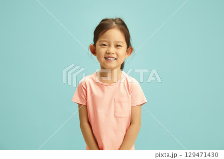 Cheerful portrait of elementary ages Asian girl radiates with a bright smile, expression joyful and happy dressed in a soft peach t-shirt standing against blue isolated background. 129471430