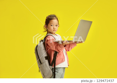 Little Asian Kid with Laptop and School Backpack stands over yellow isolated background 129471437