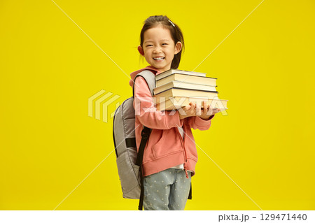 Happy chinese girl preparing going to the first grade, holds many different books in her hands and having backpack on shoulders posing on yellow isolated. Children education in preschool concept. 129471440