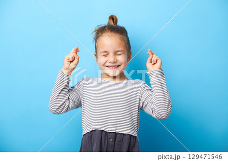 Joyful little girl clenches teeth, raises fingers crossed, makes desirable wish, squeezed her eyes shut, waits for good news Joyful little girl clenches teeth, raises fingers crossed, makes desirable wish, squeezed her eyes shut, waits for good news 129471546