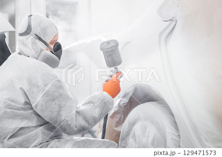 Body painter with airgun spraying white paint on a automobile in car maintenance service paint room. 129471573
