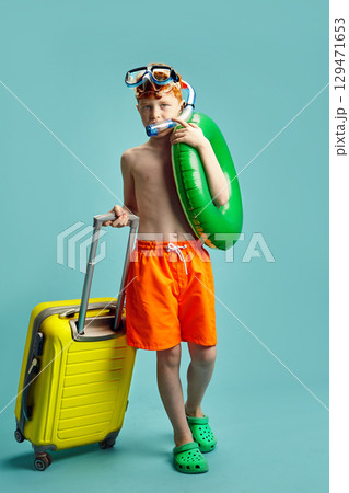 Vertical Shot of Red Headed Child Boy in Orange Swimming Trunks, with Inflatable Ring Around Himself, Having Underwater Mask and Snorkel Holding Travel Suitcase Standing on Blue Isolated Background.  129471653