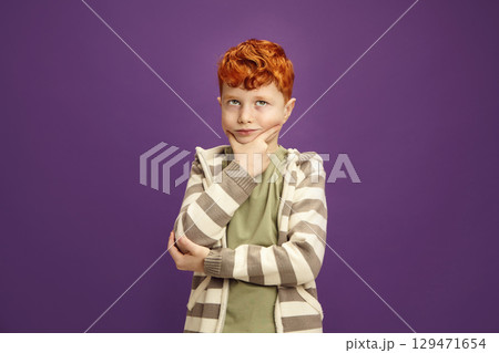 Pensive Red Headed Little Boy Around 7-8 years Puzzled Standing Keeping Hand to Cheek with Thoughtful Face Expression Looking Upwards, Wearing in Casual Clothes Standing on Isolated Violet Background 129471654