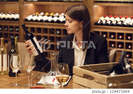 Focused female wine expert during tasting wearing in classic clothes holding bottle of red wine and reads label with characteristics of wine, the year of manufacture of the alcoholic beverage. 129471664