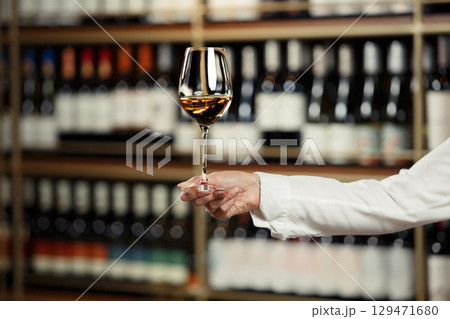 Glass of wine filled with white wine in female hand on cellar and bottles shelf background, close up with a free copy space for your text. Glass of wine filled with white wine in female hand on cellar and bottles shelf background, close up with a free copy space for your text. 129471680