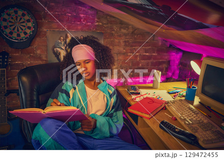 Teen girl reading book in retro room with vintage computer 129472455