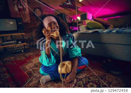 Smiling teen girl in retro tracksuit sitting on floor with vintage phone 129472459