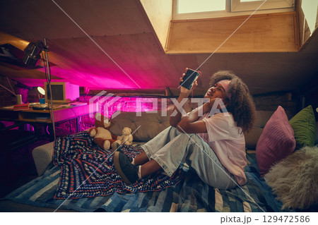 Thoughtful teenage girl listens to music on cassette player in colorful attic room. 129472586