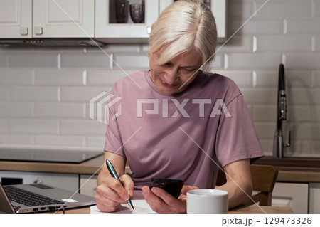 Focused Senior Woman Engaged in Multi-tasking with Phone and Notepad at Kitchen Table, Balancing Work and Daily Life Activities Focused Senior Woman Engaged in Multi-tasking with Phone and Notepad at Kitchen Table, Balancing Work and Daily Life Activities 129473326