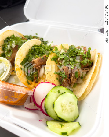 Three tacos topped with herbs and onions placed in a white takeout tray along with radish and cucumber slices. The image is bright and appetizing. Three tacos topped with herbs and onions placed in a white takeout tray along with radish and cucumber slices. The image is bright and appetizing. 129473490