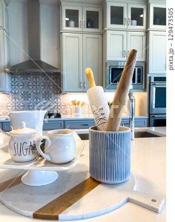Close-up of a modern kitchen counter styled with ceramic mugs, a striped vase holding utensils, and a decorative tray. Light-toned cabinets and stainless appliances appear in the background. 129473505