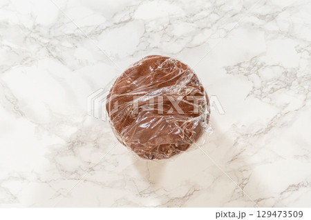 Close-up of pale gingerbread dough wrapped in plastic on a marble countertop for Pale Gingerbread Cookies with Subtle Spice Flavor. The dough is smooth and evenly shaped, ready for chilling. Close-up of pale gingerbread dough wrapped in plastic on a marble countertop for Pale Gingerbread Cookies with Subtle Spice Flavor. The dough is smooth and evenly shaped, ready for chilling. 129473509