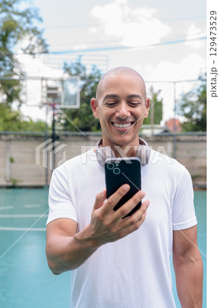 Latin man using smartphone on outdoor basketball court, urban sports fitness lifestyle 129473529