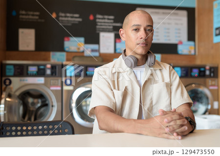 Bald Latin man sitting in laundromat with washing machines in background Bald Latin man sitting in laundromat with washing machines in background 129473550