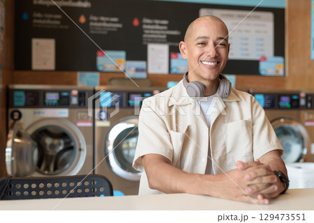 Bald Latin man sitting in laundromat with washing machines in background 129473551