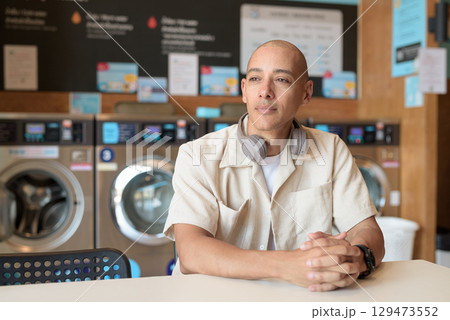 Bald Latin man sitting in laundromat with washing machines in background 129473552