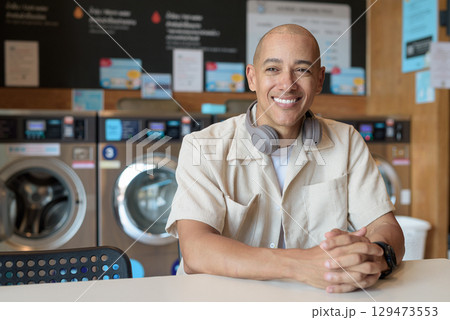 Bald Latin man sitting in laundromat with washing machines in background 129473553