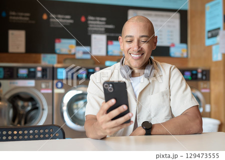 Bald Latin man using smartphone in laundromat with washing machines in background Bald Latin man using smartphone in laundromat with washing machines in background 129473555
