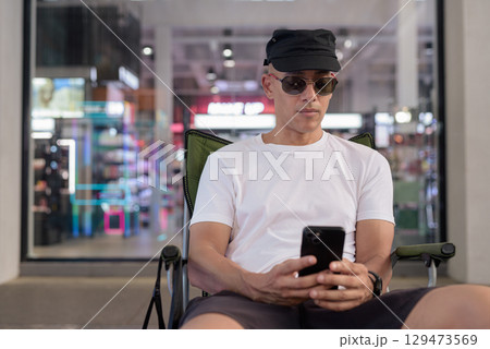 Latino man sitting outdoors in coffee shop using phone wearing sunglasses and cap 129473569