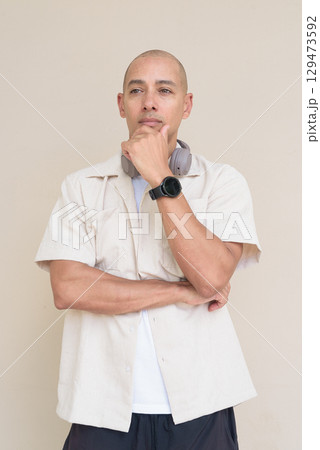 Bald Latin man in casual shirt with headphones around neck thinking with hand on chin Bald Latin man in casual shirt with headphones around neck thinking with hand on chin 129473592