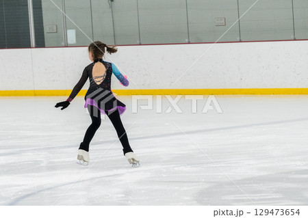 Young girl perfecting her figure skating routine while wearing her competition dress at an indoor ice rink. Young girl perfecting her figure skating routine while wearing her competition dress at an indoor ice rink. 129473654