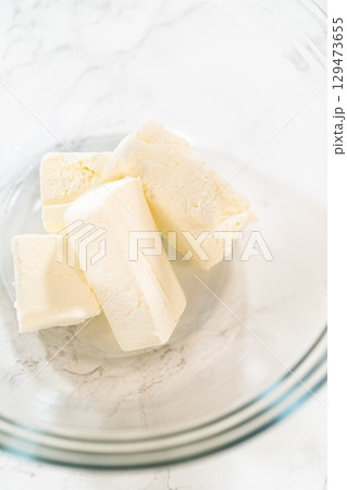 A close-up of softened butter cut into cubes and placed in a glass mixing bowl on a marble countertop. The point of view provides a clear look at the creamy texture, making it ideal for baking and 129473655