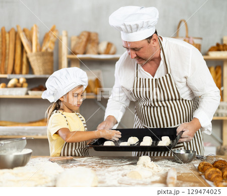 Little girl in striped apron and white chefs hat near kitchen table puts croissants on baking sheet Little girl in striped apron and white chefs hat near kitchen table puts croissants on baking sheet 129474025