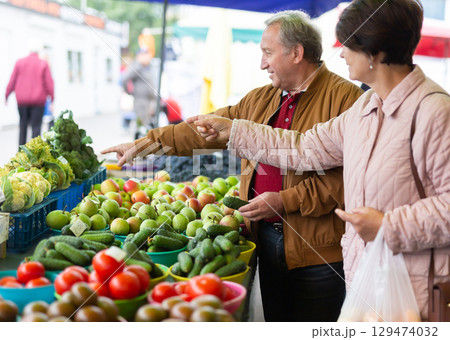 Positive mature man and woman choosing organic cucumbers near vegetable stand at local grocery market 129474032