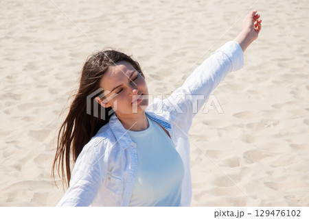 Young woman with long brown hair standing on sandy beach with eyes closed and arms raised, enjoying the sunlight, symbolizing freedom, happiness, and summer vacation Young woman with long brown hair standing on sandy beach with eyes closed and arms raised, enjoying the sunlight, symbolizing freedom, happiness, and summer vacation 129476102