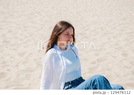 Young woman with long brown hair sitting on sandy beach under bright sunlight, looking into the distance, symbolizing summer vacation, freedom, and mindful lifestyle Young woman with long brown hair sitting on sandy beach under bright sunlight, looking into the distance, symbolizing summer vacation, freedom, and mindful lifestyle 129476112