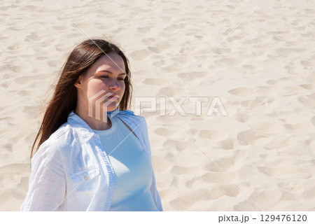 Young woman with long brown hair standing on sandy beach under bright sunlight, looking into the distance, symbolizing summer vacation, freedom, and mindful living Young woman with long brown hair standing on sandy beach under bright sunlight, looking into the distance, symbolizing summer vacation, freedom, and mindful living 129476120