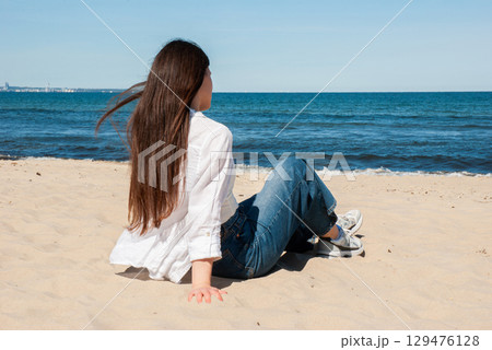 Young woman with long brown hair sitting on sandy beach and looking at the sea on a sunny day, symbolizing travel, freedom, and peaceful summer vacation 129476128