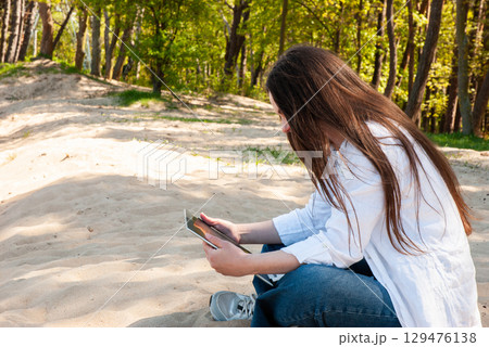 Young woman with long brown hair sitting on sandy ground in a sunny park, using a tablet device, symbolizing remote work, e-learning, and digital nomad lifestyle Young woman with long brown hair sitting on sandy ground in a sunny park, using a tablet device, symbolizing remote work, e-learning, and digital nomad lifestyle 129476138