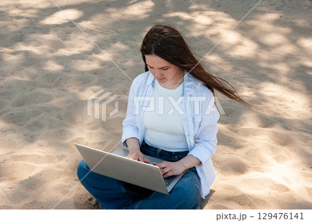 Young woman with long brown hair sitting cross legged on sandy ground in a sunny park, working on a laptop, symbolizing remote work, freelancing, and digital nomad lifestyle 129476141