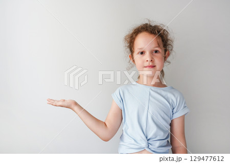 A child girl stands against a plain background, holding one hand out with an open palm as if presenting something 129477612
