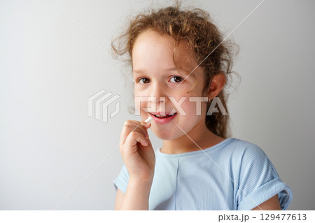 A smiling child girl in a light blue shirt holds a white tablet near her mouth, appearing ready to take it A smiling child girl in a light blue shirt holds a white tablet near her mouth, appearing ready to take it 129477613