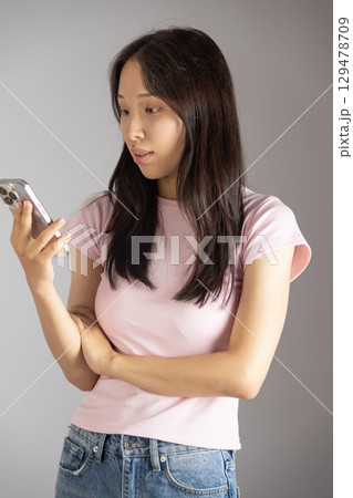 A young woman in a pink t-shirt and jeans is looking at her smartphone with a focused expression A young woman in a pink t-shirt and jeans is looking at her smartphone with a focused expression 129478709
