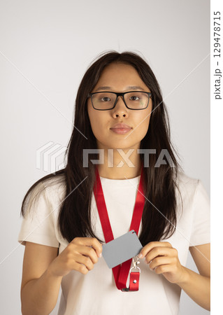 A young woman wearing glasses and a red lanyard with an ID card is holding a blank grey card in front of her 129478715